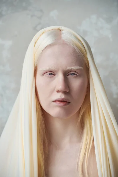 Young serene albino woman with white cover on head standing in front of camera against marble wall during photo session in studio