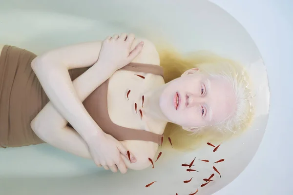 身着米黄色坦克衫的年轻沉着的白化病妇女抱着胳膊躺在装满清澈水的浴缸里 望着小鱼 — 图库照片