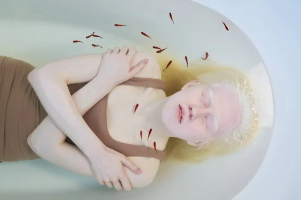 Above view of young pale woman with albinism lying in clear water in bathtub while stock of tiny fish swimming over her chest