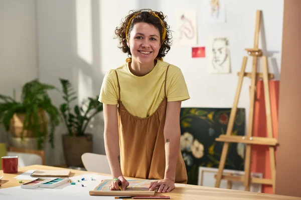 Young cheerful female artist standing by workplace with supplies for drawing in home studio against sketches on wall and easel