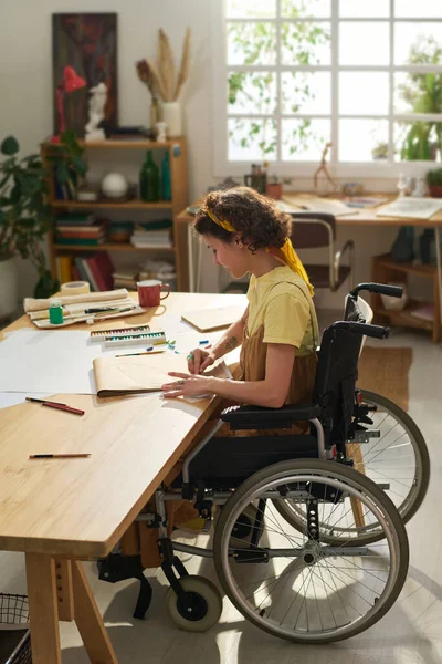 stock image Adolescent girl in wheelchair sitting by wooden table with supplies for art creation in her home studio and drawing new masterpiece