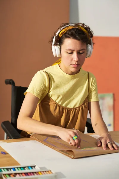 残疾年轻而严肃的女工匠 一边用耳机听音乐 一边在纸上用蜡笔画草图 — 图库照片