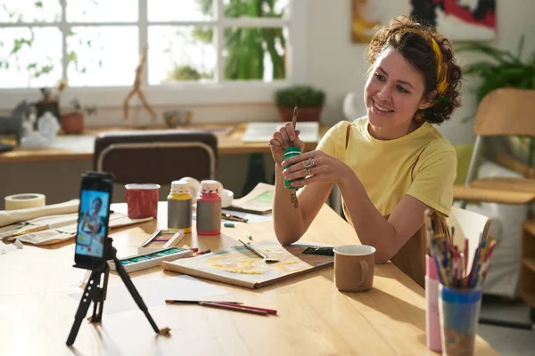 stock image Young smiling creative woman with paintbrush and gouache sitting by workplace and looking at smartphone camera during masterclass