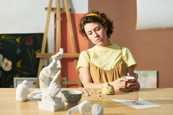 自宅のスタジオで仕事をしながらお茶を飲みながらテーブルの上で未完成の粘土作品を見る若い女性アーティスト — ストック写真