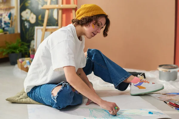 自宅のスタジオやワークショップの床に座っている間にクレヨンを使用してカジュアルウェアで若い創造的な女性や紙の上にスケッチ — ストック写真