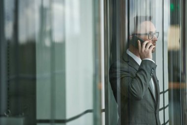Asansörde yürürken cep telefonundan konuşan zarif, koyu gri takım elbiseli ve gözlüklü, kendinden emin genç bir girişimci.