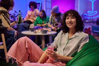 Arkadaşları arka planda dinlenirken elinde sodayla kameranın önünde oturup partiden zevk alan mutlu ergen kız.