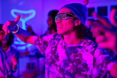 Neon gözlüklü ve günlük giysili genç adam ev partisinin keyfini çıkarırken kameranın önünde dans ediyor.