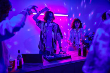 Genç erkek DJ pikapta müzik yapıyor ve dans ediyor. Evde partisinde kız arkadaşının yanında elinde bira şişesiyle dikiliyor.