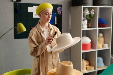 Sarı bereli genç yaratıcı kadın ve şık günlük kıyafetler moda stüdyosunda duruyor ve yeni beyaz keçeli şapkayı fırçayla temizliyor.