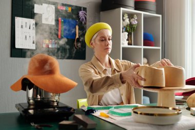 Genç bir zanaatkar atölyede masa başında otururken ve yeni eşyalar üzerinde çalışırken şapka için ahşap işyerinin üst kısmını değiştiriyor.