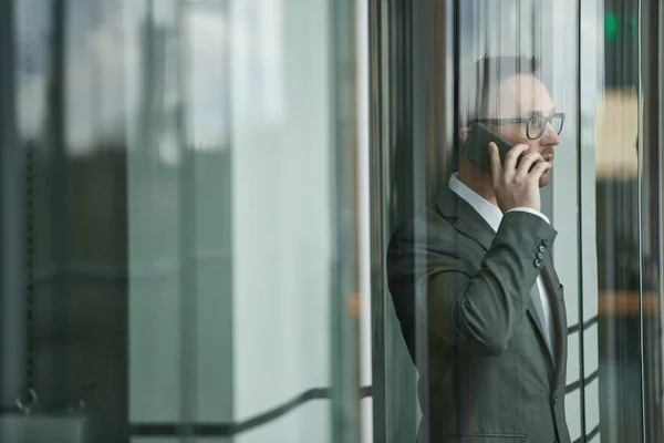 エレベーターに移動しながら携帯電話で話すエレガントなダークグレーのスーツと眼鏡で若い自信に満ちた男性起業家 — ストック写真