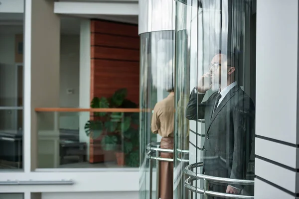 穿着西装的年轻男性经纪人在透明电梯里与同事用智能手机交谈 同时进入现代办公中心 — 图库照片