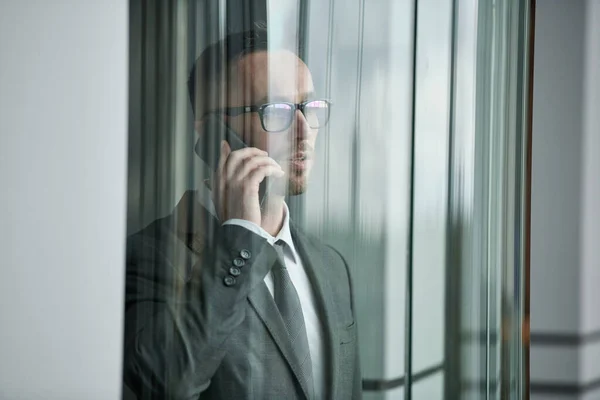 穿着正装和眼镜的年轻商人在现代办公中心的透明玻璃电梯里对着手机讲话 — 图库照片