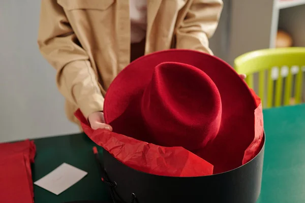 年轻的女工匠把新的深红色的毛毡帽放进黑色的圆盒子里 然后再把它包装成工艺用纸 为顾客包装 — 图库照片