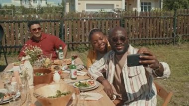 Modern Afro-Amerikan çifti yemek masasında oturuyor ve yaz günü açık hava partisinde akıllı telefon kamerasıyla selfie çekiyorlar.