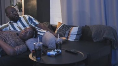 Kocasının kucağında uzanan genç bir kadının solunda. Kanepede oturan, geceleri televizyon izleyen bir çift.