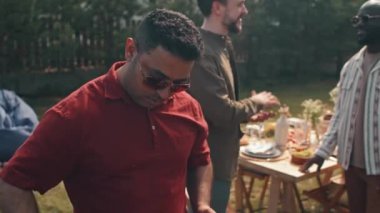 Orta Doğulu adam kırmızı tişört ve güneş gözlüğü takıyor yaz günü arka bahçe partisi için ızgaraya sosis koyuyor.