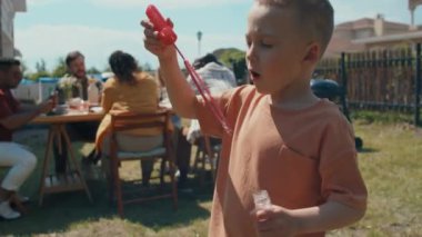 Kafkas çocuk portresi dışarıda sabun köpükleri üflüyor, bir grup yetişkin arkadaş masada oturmuş yemek yiyor ve arka planda sohbet ediyorlar.