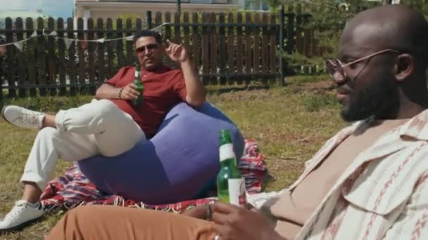 現代の黒と中東の男性は 裏庭でビールを飲みながら豆の袋の椅子に座って夏の日に話してリラックス — ストック動画