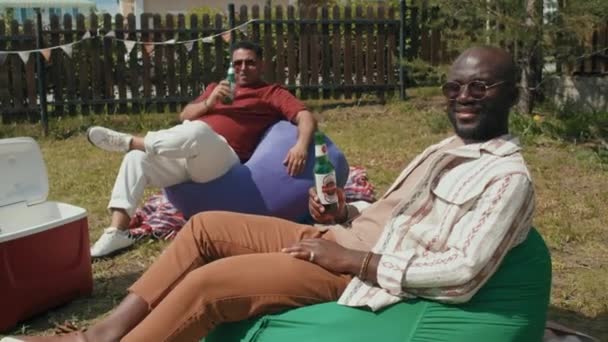 現代アフリカ系アメリカ人と中東人の男性の肖像カメラで笑顔ビールのボトルを保持裏庭で豆の袋の椅子にリラックス座って — ストック動画
