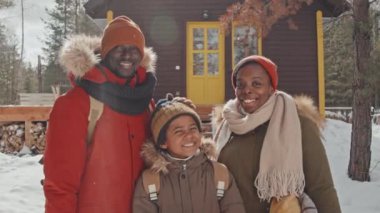 Modern Afro-Amerikan erkek, kadın ve oğullarının, kış günü kır evinin karşısında kameraya gülümseyişlerinin ağır çekimde portresi.