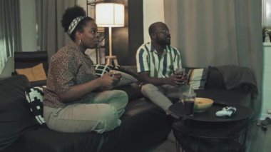 Orta uzunlukta romantik bir çift oturma odasında kanepede oturur, abur cubur yer, içki içer, kadın geceleri bilgisayar oyunu oynar.