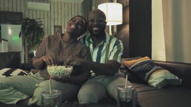 Orta uzunlukta Afro-Amerikan karı-koca kanepede dinleniyor, televizyon izliyor, patlamış mısır yiyor, konuşuyor ve gülüyor.