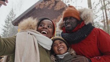 Üç kişilik şık Afro-Amerikan ailesi, karlı bir günde selfie çekerek orman köyündeki kır evlerine karşı sıcak kıyafetler giyiyorlar.