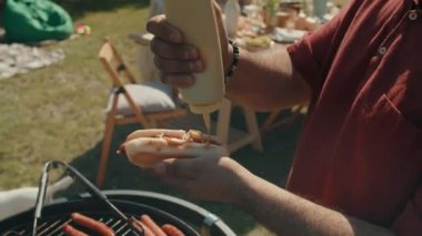 Ortadoğulu bir adam güneşli yaz gününde açık hava partisinde hardallı sosisli sandviç yapıyor.