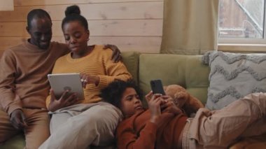 Çocuklu modern Afro-Amerikan ailesi kış günü kır evinde dinleniyor dijital tablet ve akıllı telefonla internette sörf yapıyor.