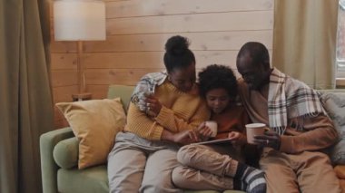 Mutlu siyah aile ve çocuk soğuk kış gününde evde birlikte rahatlıyor kanepede oturuyor, sıcak süt ve sıcak çay içiyor ve dijital tablette internette sörf yapıyor.