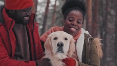 Genç Afrikalı Amerikalı eşlerin hafta sonları çam ormanlarında mutlu Golden Retriever köpeğini okşamaları yavaş bir harekettir.