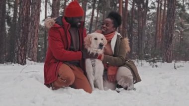 Genç Afrikalı Amerikalı eşlerin hafta sonu çam ormanlarında karda diz çöküp mutlu Golden Retriever köpeğini okşamaları.