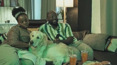 Orta uzunluktaki Afro-Amerikan kadın ve Golden Retriever köpekli bir adam oturma odasında oturmuş, akşamları televizyon izliyorlar.