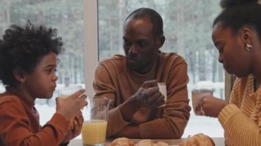 Modern Afrikalı erkek, kadın ve oğulları kış günü evde oturup taze meyve suyu ile kruvasan yiyor ve tatil planları hakkında konuşuyorlar.