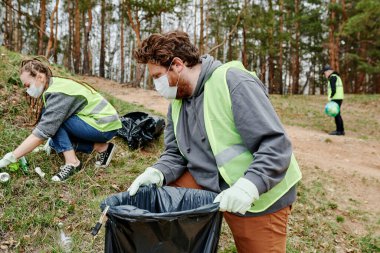Bir grup arkadaşla ormanda çöp toplarken koruyucu maske takan bir gönüllü.
