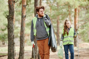 Kaşlarını çatmış gönüllü ormanda çöp torbası taşıyor.