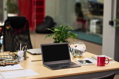 Siyah ekranlı dizüstü bilgisayar, çiçek saksısında yeşil bitki, kırmızı bardak, tablet, su ve diğer şeylerle dolu küresel akvaryum.