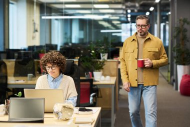 Kırmızı kahve fincanı olan olgun bir iş adamı genç esmer kadın iş arkadaşının yanından geçerken ağ sırasında dizüstü bilgisayarın önünde oturuyor.