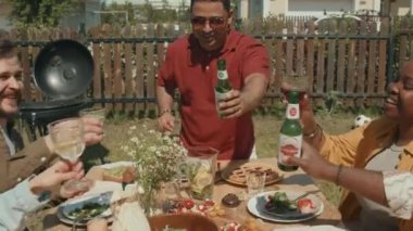 Etnik çeşitlilikteki en iyi arkadaşlar yemek masasında oturup bardakları tokuşturarak ve şişeleri içerek güneşli yaz gününde bir şeyler kutluyorlar.