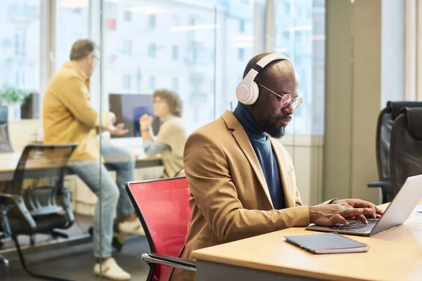 Poważny Afro Amerykański Pracownik Słuchawkami Siedzącymi Przy Biurku Przed Laptopem — Zdjęcie stockowe