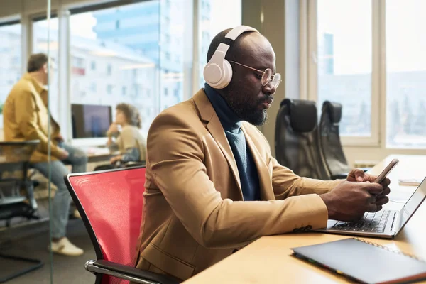 stock image Serious young businessman in headphones choosing new track from playlist in mobile phone while working in front of laptop