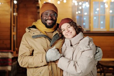 Sıcak kışlık giysili mutlu bir Afrikalı genç adam güzel karısını kucaklarken ikisi de kulübeye karşı kameraya bakıyor.