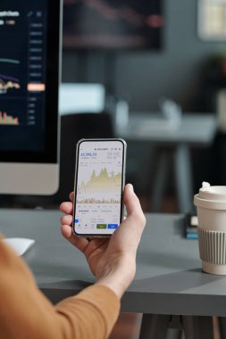 Genç borsa analistinin elinde iş yerinin yanında otururken ekranda finansal istatistiklerle akıllı telefon var.