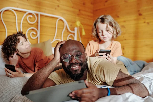 タブレットを持っている男と彼の妻と息子のスマートフォンでテキストメッセージながら ベッドの中でモバイルガジェットを使用して若い安らかな異文化間の家族 — ストック写真