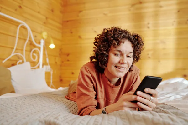 곱슬머리의 여자가 핸드폰을 사용하여 아침에 잠자리에서 긴장을 문자를 보내는 — 스톡 사진