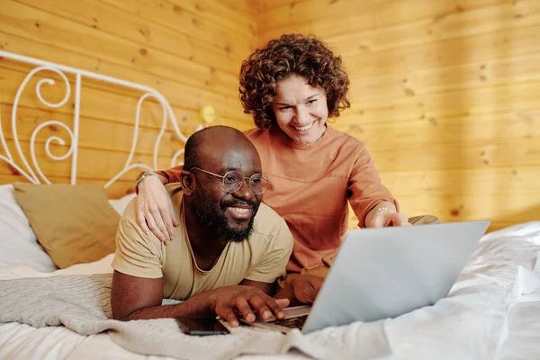 Молода Усміхнена Жінка Обіймає Чоловіка Вказує Екран Ноутбука Одночасно Відпочиваючи — стокове фото