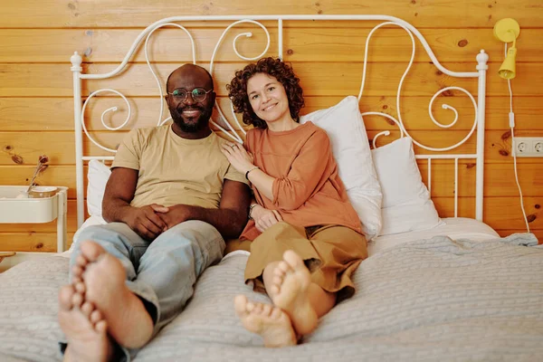 身穿睡衣 面带微笑的年轻女子坐在非洲裔美国人丈夫旁边 一边放松 一边享受周末 一边看着镜头 — 图库照片