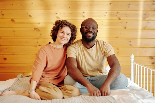 幸せな若いです異文化間のカップルでホームウェアで座っている上のダブルベッドと見てカメラに対して木製の壁のベッドルームでコテージ — ストック写真